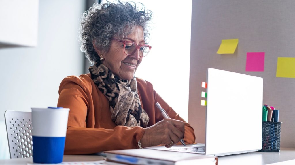 older woman sat at desk with laptop working, has a contented smile on her face