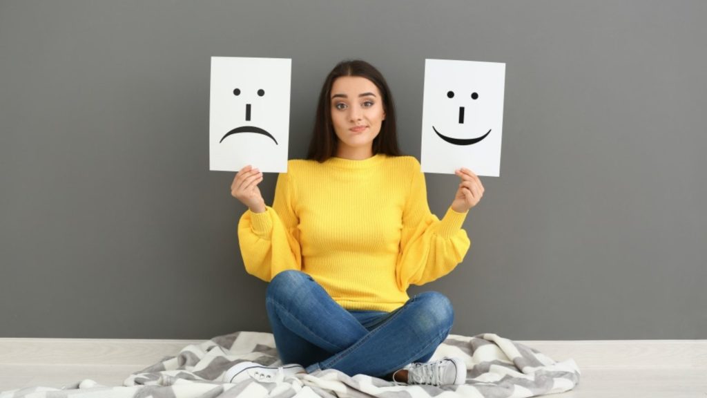 woman sat crossed legged holding a sad smile card in one had and a smiley face in the other. Her facial expression is of someone who may not feel she is good enough
