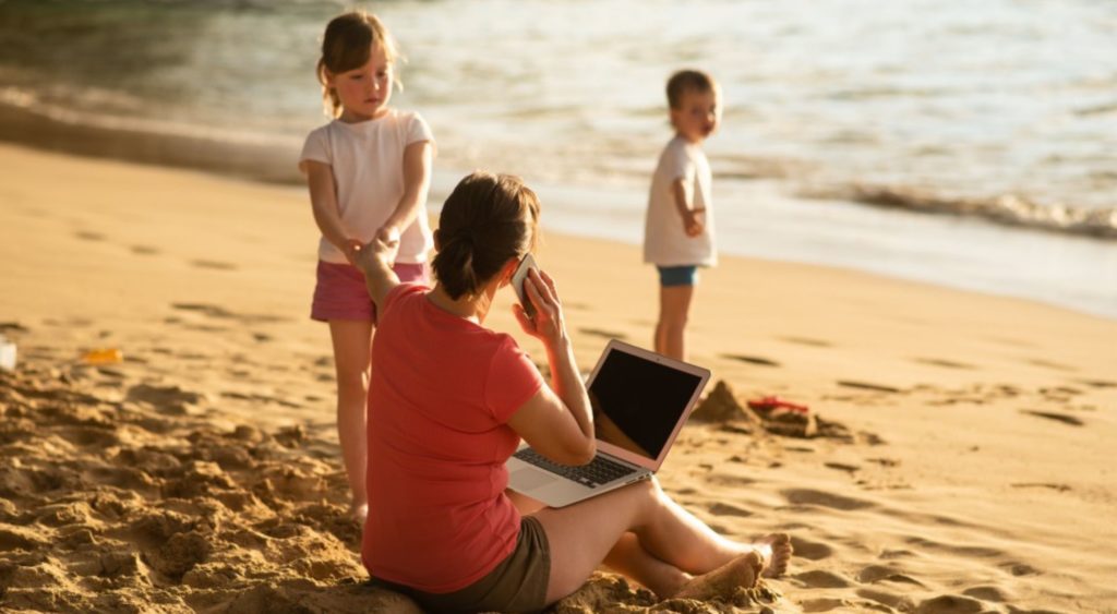 woman on beach with her 2 children, is working on her laptop. one child is trying to distract her from working, pulling her hand (all work and no play leaves you stressed and headed for burnout, so disconnect from work on your summer break)